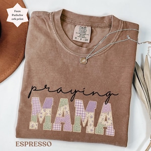 Puede incluir: Camiseta color espresso con las palabras "praying MAMA" en un diseño de patchwork. La camiseta tiene cuello redondo y mangas cortas. Un collar plateado con un colgante está drapeado sobre la camiseta. La camiseta está etiquetada como "Comfort Colors".