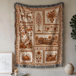 May include: A rectangular woven throw blanket featuring a multi-panel design in shades of orange, brown, and cream. The panels display autumn-themed illustrations including deer, a tree, a floral vase, a truck with pumpkins, and a cornucopia.