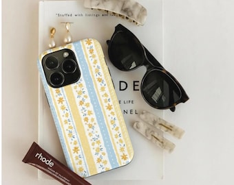 Op de afbeelding: Een smartphonehoesje met een verticaal streeppatroon in geel en lichtblauw met kleine witte bloemaccenten. Het hoesje wordt omringd door een marmeren haarklem, een zwarte zonnebril, twee kleine haarspeldjes en een bruine tube met het opschrift rhode.