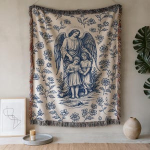 May include: Cream-colored woven throw blanket with a blue angel illustration and floral pattern. The image depicts an angel with two children, surrounded by flowers. The blanket has a fringed edge and is displayed on a wall.