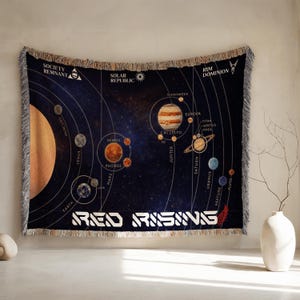 Una manta tejida con diseño del sistema solar y texto blanco cuelga estirada en una pared junto a un jarrón.