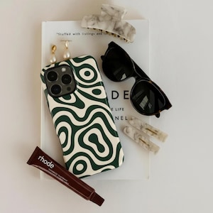 Puede incluir: Un surtido de accesorios que incluye una funda de teléfono con un diseño de remolino verde y blanco, gafas de sol, pinzas para el pelo, pendientes y un tubo de producto labial. Los artículos están colocados sobre un libro.