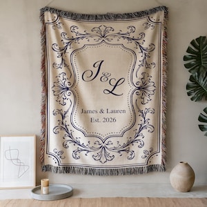 Puede incluir: Una manta tejida de color crema con un borde de filigrana ornamentado en azul oscuro y un monograma central con las letras J & L. Debajo del monograma, el texto dice James & Lauren Est. 2026. La manta tiene bordes con flecos en todos los lados.