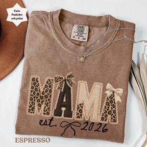Peut inclure: T-shirt couleur expresso avec le mot "MAMA" en imprimé patchwork. Les lettres ont un motif léopard et sont agrémentées de nœuds. Le texte "est. 2026" est en dessous du mot "MAMA". Un collier argenté est drapé sur le t-shirt.