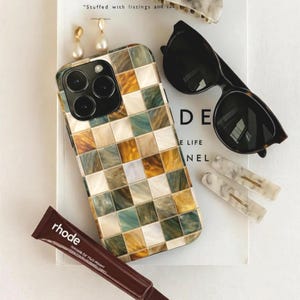 Puede incluir: Un surtido de accesorios que incluyen una funda para teléfono, gafas de sol, pinzas para el pelo, pendientes y un tubo de producto labial. La funda del teléfono tiene un patrón de cuadros en tonos verdes, dorados y marrones. Las gafas de sol son negras.