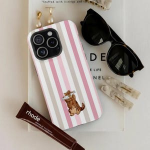 Puede incluir: Un surtido de accesorios que incluyen una funda de teléfono con diseño de rayas rosas y blancas y una ilustración de gato con peces. También se muestran gafas de sol, pinzas para el pelo, pendientes y un tubo de producto cosmético marrón.