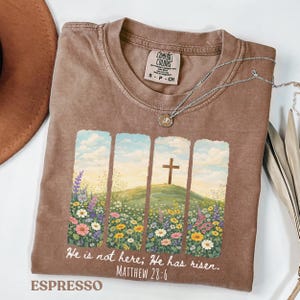 Puede incluir: Camiseta color espresso con un gráfico floral y una cruz. El diseño presenta una cruz sobre una colina con un campo de flores coloridas. El texto "He is not here; He has risen. Matthew 28:6" está impreso debajo. Un collar plateado está sobre la camiseta.