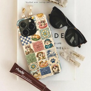 Puede incluir: Una colección de accesorios que incluye una funda de teléfono con diseño de azulejos de gatos, gafas de sol, pinzas para el pelo, pendientes y un tubo de producto labial. La funda presenta ilustraciones de gatos.