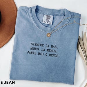 Puede incluir: Una camiseta azul vaquero Comfort Colors con el texto "SIEMPRE LA MÁS, NUNCA LA MENOS, JAMÁS MÁS O MENOS." Un collar dorado con un colgante descansa sobre la camiseta. Un sombrero marrón y una planta seca están en el fondo.