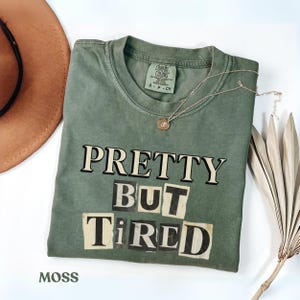 Puede incluir: Camiseta verde musgo con las palabras "PRETTY BUT TIRED" en un estilo recortado. Un collar dorado y un sombrero marrón están al lado de la camiseta. La camiseta está hecha de un material suave y tiene un ajuste cómodo.