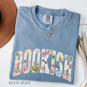 Puede incluir: Una camiseta azul vaquera Comfort Colors con la palabra "BOOKISH" en un diseño blanco con temática playera. El diseño incluye un sol, palmeras y una silla de playa. También se ve un collar dorado con un colgante.