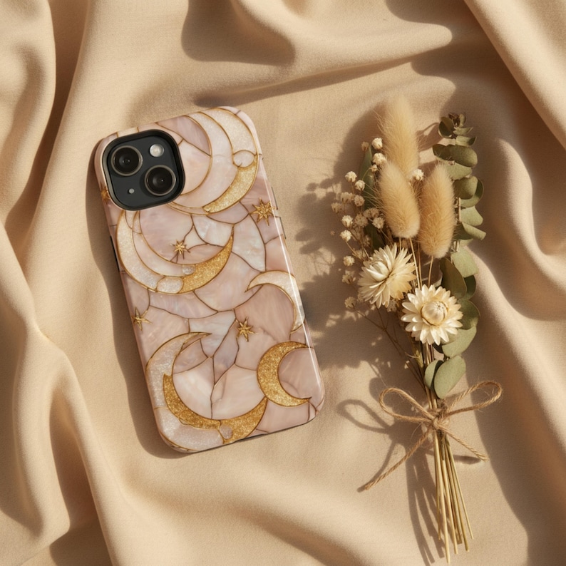 Fundador de teléfono con diseño de lunas doradas sobre fondo rosa marmolado, colocado sobre tela beige junto a un pequeño ramo de flores secas.
