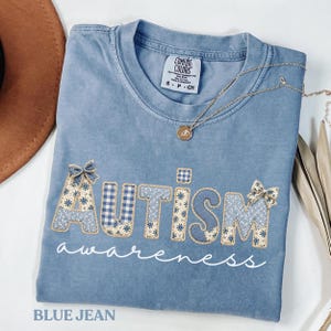 Puede incluir: Una camiseta azul vaquera con la palabra "AUTISMO" aplicada en tela de retazos, con un lazo a cada lado. La palabra "awareness" está escrita debajo en cursiva. Un collar de oro con un colgante descansa sobre la camisa.