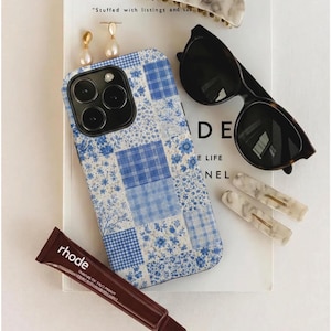 Puede incluir: Una colección de accesorios que incluye una funda de teléfono con estampado floral azul, gafas de sol, pinzas para el pelo de mármol y pendientes de perlas. También hay un tubo de tratamiento labial rhode. Los artículos están colocados sobre un libro con texto.