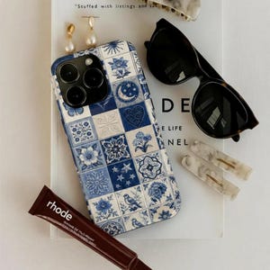 Puede incluir: Una colección de accesorios que incluye una funda de teléfono con estampado azul y blanco, gafas de sol, pinzas para el pelo, pendientes y un tubo de producto labial. La funda del teléfono presenta un diseño de azulejos florales y celestiales.
