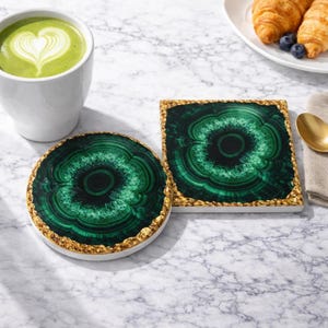 Puede incluir: Dos posavasos decorativos con diseño de malaquita. Uno es redondo, el otro cuadrado. Ambos tienen un borde dorado. Una taza blanca con una bebida verde y un diseño de corazón está en el fondo.