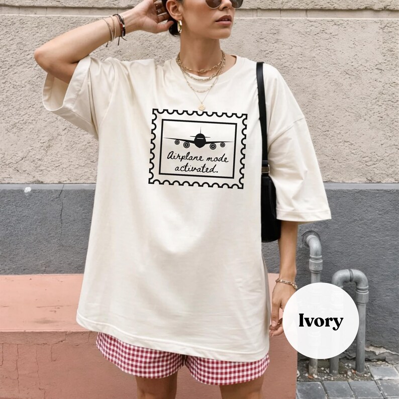 Puede incluir: Camiseta de color marfil con un gr&aacute;fico negro de un avi&oacute;n dentro de un dise&ntilde;o de sello postal. El texto "Airplane mode activated" est&aacute; debajo. La modelo lleva pantalones cortos a cuadros rojos y blancos.