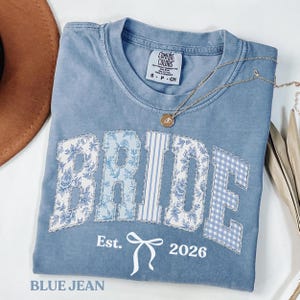 Puede incluir: Una camiseta azul vaquero con la palabra "BRIDE" en un diseño de patchwork. Las letras tienen patrones florales, de rayas y cuadros. Debajo de la palabra "BRIDE" está "Est. 2026" con un gráfico de lazo. Un collar dorado con un colgante está sobre la camiseta.