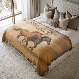 Puede incluir: Una manta tejida con un diseño de dos caballos corriendo en tonos marrones y beige. La manta tiene un borde con flecos y el texto "Olivia & Theo Est. 2008" en escritura negra. Se muestra sobre una cama con almohadas.