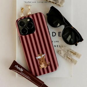 Puede incluir: Colección de accesorios: funda de teléfono, gafas de sol, pinzas para el pelo y pendientes. La funda es rosa con rayas verticales burdeos y un dibujo de gato. Las gafas son negras con montura carey.