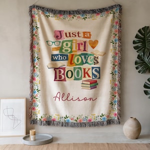 Puede incluir: Una manta crema con un borde floral y flecos. La manta presenta el texto "Just a girl who loves BOOKS" en letras de colores, con el nombre "Allison" debajo. El diseño incluye gafas, un corazón y libros abiertos.