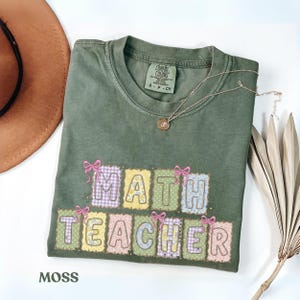Puede incluir: Una camiseta verde oliva con las palabras "MATH TEACHER" en un diseño de patchwork colorido. La camiseta tiene cuello redondo y mangas cortas. También se ve un collar dorado y un sombrero marrón.