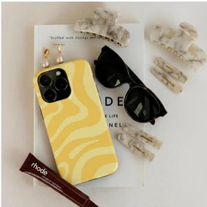 Puede incluir: Una colección de accesorios que incluye una funda de teléfono amarilla y crema con diseño de remolino, gafas de sol negras, pinzas para el pelo con efecto mármol, pendientes dorados y un tubo de producto labial marrón. Los artículos están sobre un libro con el texto "Stuffed with listings and..."