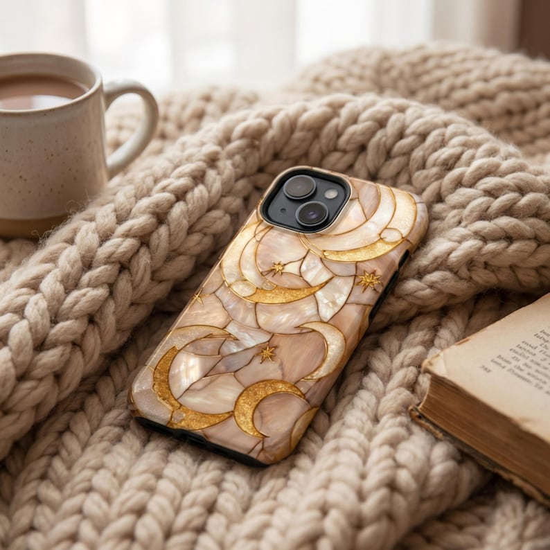 Funda de teléfono con patrón de fases lunares doradas reposando sobre una manta de punto grueso, junto a una taza y un libro abierto.