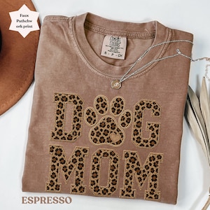 May include: Espresso-coloured t-shirt featuring the words "DOG MOM" in a leopard print design. The shirt has a crew neck and short sleeves. A silver necklace with a pendant is draped across the shirt.
