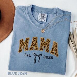 Puede incluir: Una camiseta azul vaquera con la palabra "MAMA" en letras con estampado de leopardo. Debajo de la palabra "MAMA" está el texto "Est. 2026" con un gráfico de lazo negro. Un collar dorado con un colgante está drapeado sobre la camisa.