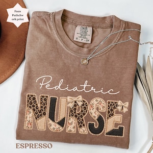 Puede incluir: Camiseta color espresso con las palabras "Pediatric Nurse" en escritura blanca y letras patchwork. Las letras presentan estampado de leopardo, patrones florales y detalles en negro. Un collar plateado con un colgante está sobre la camiseta.
