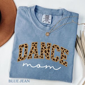 Könnte beinhalten: Ein blaues Jeans-T-Shirt mit den Worten "DANCE mom" auf der Vorderseite gestickt. Das Wort "DANCE" ist in einem Leopardenmuster gehalten. Eine goldene Halskette mit Anhänger liegt auf dem Shirt.