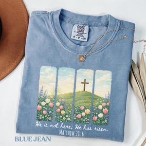 Puede incluir: Camiseta azul vaquero con un gráfico de una cruz sobre una colina, rodeada de flores. El texto "He is not here; He has risen. Matthew 28:6" está impreso debajo. Un collar dorado está sobre la camiseta.