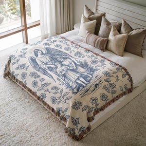 May include: A cream-colored woven throw blanket featuring a blue illustration of an angel with two children. The blanket has a floral border and fringed edges. The throw is displayed on a bed with decorative pillows.
