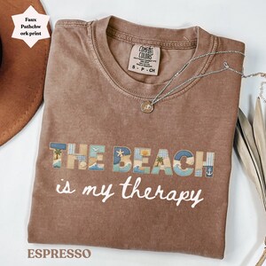 Puede incluir: Camiseta color espresso con la frase "THE BEACH is my therapy" en escritura blanca. Las palabras "THE BEACH" están en un diseño de patchwork con imágenes de temática playera. Un collar plateado con un colgante descansa sobre la camiseta.