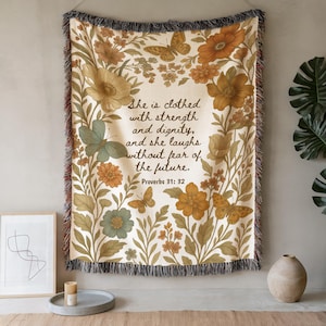 Pode incluir: Uma manta tecida com um design floral e borboletas em tons de creme, laranja e verde. O texto diz: "She is clothed with strength and dignity, and she laughs without fear of the future. Proverbs 31:32".