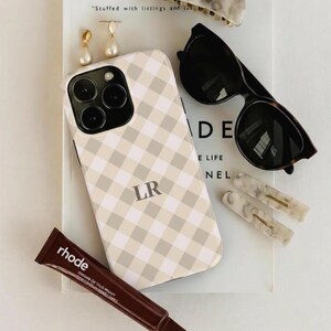 Puede incluir: Una colección de accesorios que incluye una funda de teléfono con un patrón de cuadros beige y blanco y las iniciales "LR", gafas de sol, pinzas para el pelo, pendientes y un tubo de tratamiento labial rhode. Los artículos están colocados sobre un libro.