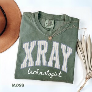 Puede incluir: Una camiseta verde oliva con la palabra "XRAY" en una fuente de estilo universitario, con rayas verticales azules y la palabra "technologist" debajo. Un collar dorado y un sombrero marrón están al lado de la camisa. La camisa está etiquetada como "MOSS".