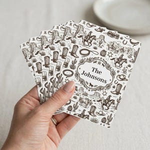 Puede incluir: Una mano sostiene una pila de tarjetas blancas con un diseño de temática occidental en tonos sepia. El diseño incluye vaqueros a caballo, cactus, botas de vaquero y el texto "The Johnsons" en un marco decorativo. Un plato blanco está en el fondo.