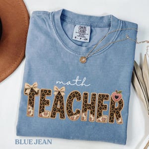 Puede incluir: Una camiseta azul vaquera con las palabras "math TEACHER" en una fuente decorativa. La palabra "TEACHER" está en un estampado de leopardo con lazos y un gráfico de manzana. También se ven un collar dorado y un sombrero marrón.
