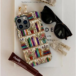 Puede incluir: Un surtido de accesorios que incluyen una funda de teléfono con diseño de estantería, gafas de sol, pinzas para el pelo, pendientes y un tubo de tratamiento labial Rhode. La funda del teléfono presenta libros coloridos y plantas en macetas.