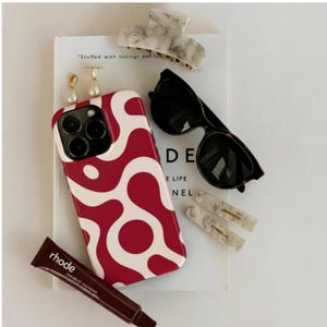 Puede incluir: Una colección de accesorios que incluye una funda de teléfono con un diseño abstracto rojo y blanco, gafas de sol, pinzas para el pelo, pendientes y un tubo de producto labial. Los artículos están colocados sobre un libro.