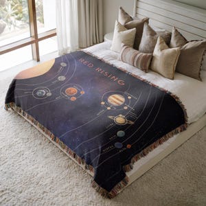 Puede incluir: Una manta azul oscuro con un diseño del sistema solar, que muestra planetas y cuerpos celestes. La manta tiene el texto "RED RISING" y un borde con flecos. Se muestra sobre una cama con almohadas.