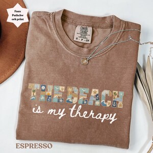 Puede incluir: Camiseta marrón con la frase "THE BEACH is my therapy" en escritura blanca. Las palabras "THE BEACH" están en un diseño de patchwork con imágenes de temática playera. Un collar plateado con un colgante está sobre la camiseta.