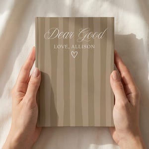 Puede incluir: Un libro de tapa dura con una cubierta de rayas verticales en color topo y beige. La portada presenta el texto blanco "Dear Good" en letra cursiva, seguido de "LOVE, ALLISON" en letra sans-serif y un pequeño corazón blanco.