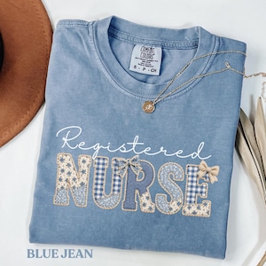 Puede incluir: Una camiseta azul vaquera con las palabras "Registered Nurse" en escritura blanca. La palabra "NURSE" está en un diseño de patchwork con estampados florales y de cuadros. También se ven un collar dorado y un sombrero marrón.