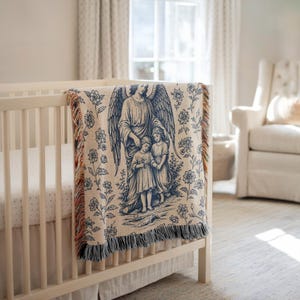 May include: A cream-colored baby blanket with a blue angel and children design, draped over a white crib. The blanket features a detailed illustration of an angel with outstretched wings, protecting two children. Floral patterns surround the central image, with fringed edges.