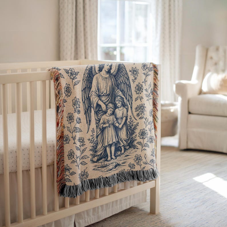 May include: A cream-colored baby blanket with a blue angel and children design, draped over a white crib. The blanket features a detailed illustration of an angel with outstretched wings, protecting two children. Floral patterns surround the central image, with fringed edges.