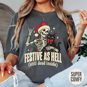 May include: A charcoal gray t-shirt featuring a graphic of a skeleton wearing a Santa hat and making a rock-and-roll hand gesture. The skeleton holds a black cat with a red bow. Text reads: FESTIVE AS HELL (still dead inside).