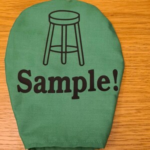 Può includere: Una sacca di stoffa verde con un contorno nero di uno sgabello e la scritta "Sample!" stampata su di essa.